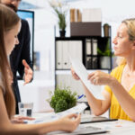Kobieta prezentuje dokument innej kobiecie, co symbolizuje poszukiwanie pracy.
