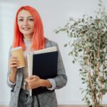 Kobieta uśmiechająca się, trzymająca szklankę piwa w pomieszczeniu z rośliną doniczkową, symbolizująca relaks po poszukiwaniach pracy.