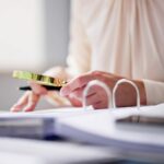Osoba pisząca w książce przy biurku, otoczona artykułami biurowymi, doskonali swoje umiejętności w poszukiwaniu pracy.