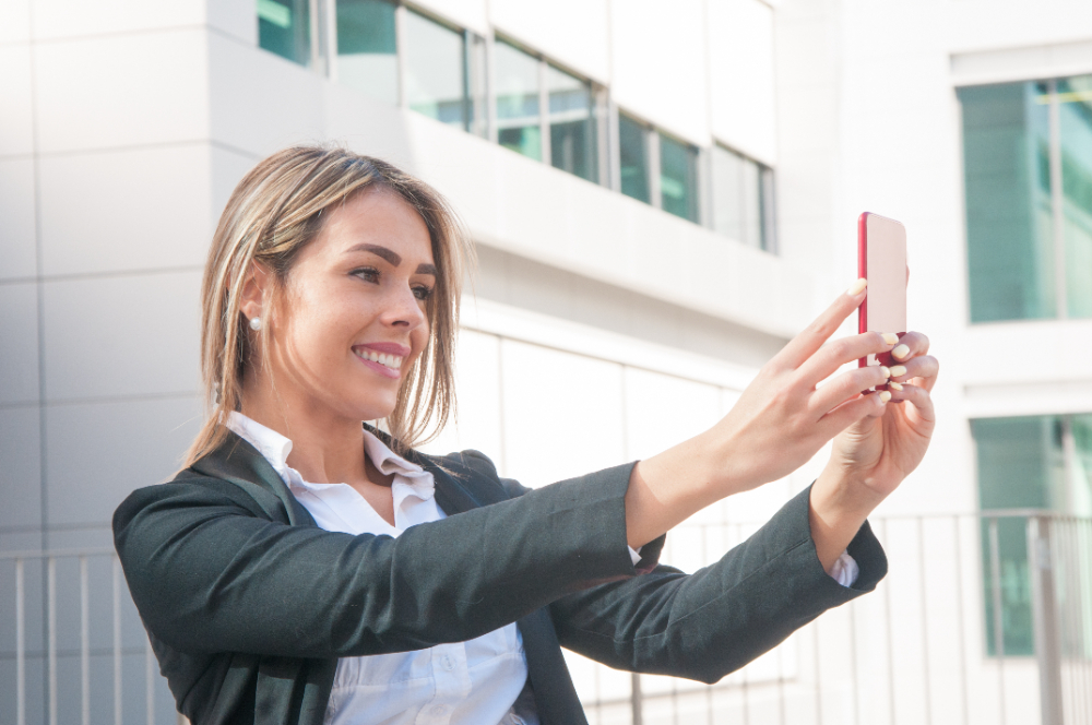 Dlaczego nie dostaję zaproszeń na rozmowę kwalifikacyjną Kobieta robiąca selfie przed budynkiem, uśmiechnięta i ubrana elegancko, idealna do promocji w poszukiwaniu pracy.