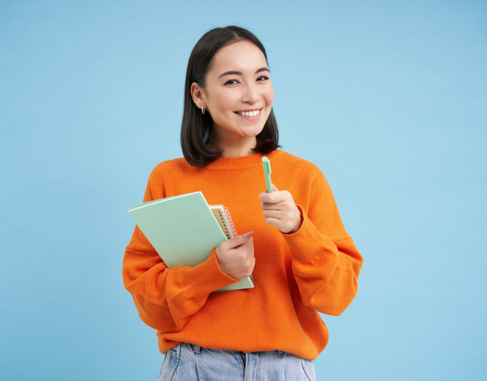 Kobieta trzymająca tablet, uśmiechnięta, w pomarańczowym ubraniu, na tle nieba, symbolizująca poszukiwania pracy. CV na staż
